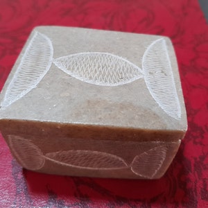 Op de afbeelding: Een kleine, vierkante, grijze stenen doos met een gesneden ontwerp van overlappende ovalen. De doos heeft een scharnierend deksel en is gemaakt van glad, gepolijst steen.
