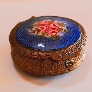 Puede incluir: Una pequeña caja de joyas redonda de color dorado con un diseño ornamentado. La tapa está decorada con un fondo de esmalte azul y un diseño floral pintado a mano con rosas rosas.