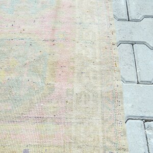 May include: A section of a vintage rug with a faded, pastel color palette. The rug features a mix of pink, blue, and yellow hues, with a textured surface. The edge of the rug is next to a section of gray paving stones.
