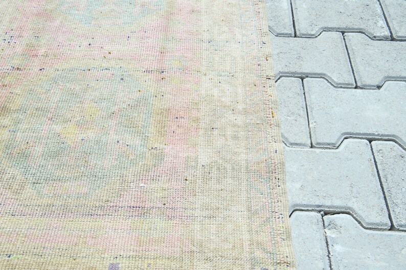 May include: A section of a vintage rug with a faded, pastel color palette. The rug features a mix of pink, blue, and yellow hues, with a textured surface. The edge of the rug is next to a section of gray paving stones.