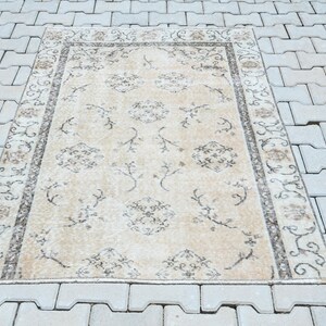 May include: Rectangular beige rug with a detailed floral pattern and a dark border. The rug features a central design of repeating floral motifs and is placed on a gray tiled surface. The rug's design is symmetrical.