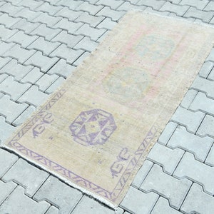 May include: A rectangular, vintage-style rug with a faded, beige background. It features geometric patterns in purple and pink, with a fringed edge. The rug is placed on a gray, interlocking paver surface.