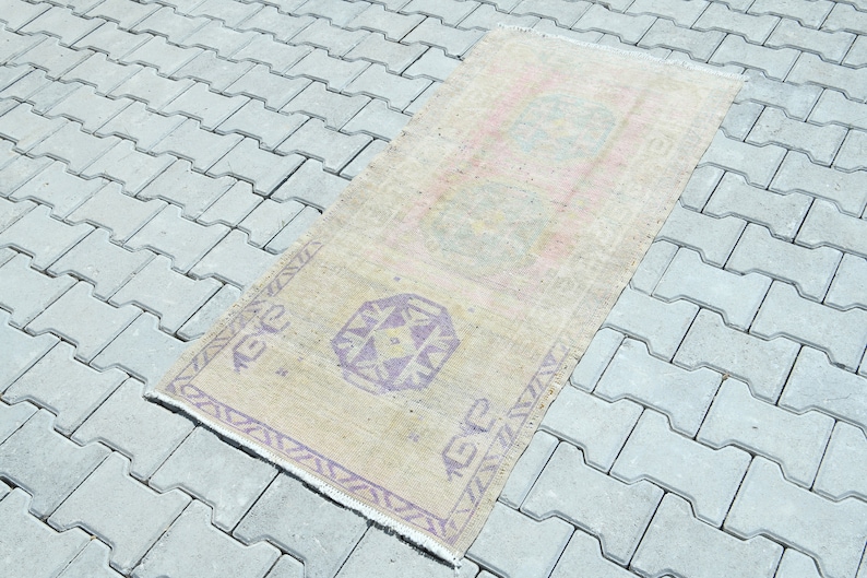 May include: A rectangular, vintage-style rug with a faded, beige background. It features geometric patterns in purple and pink, with a fringed edge. The rug is placed on a gray, interlocking paver surface.