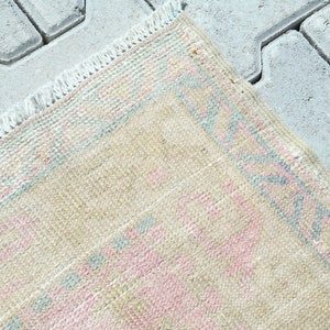 May include: Close-up of a beige rug with a pink and blue geometric pattern. The rug has a fringed edge and is placed on a gray, textured surface. The rug's design features repeating shapes.