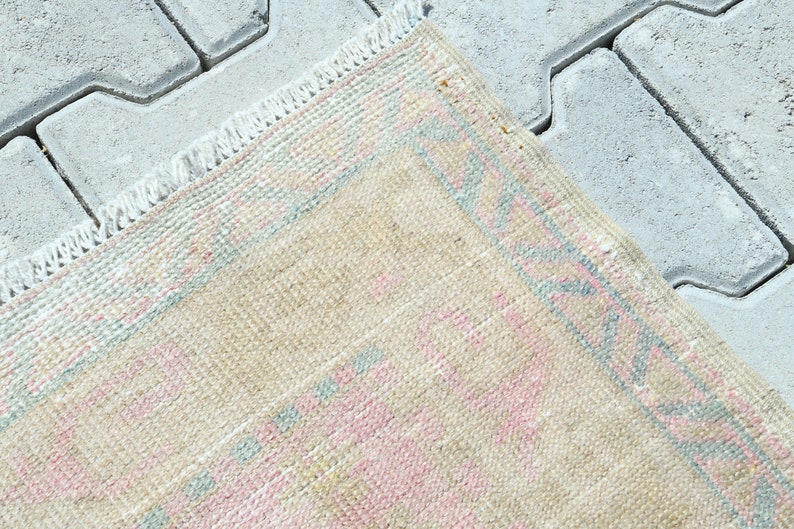 May include: Close-up of a beige rug with a pink and blue geometric pattern. The rug has a fringed edge and is placed on a gray, textured surface. The rug's design features repeating shapes.