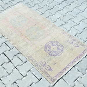 May include: A rectangular, beige rug with a faded, vintage appearance. The rug features repeating geometric patterns in shades of purple and pink. The rug is placed on a gray, concrete surface.