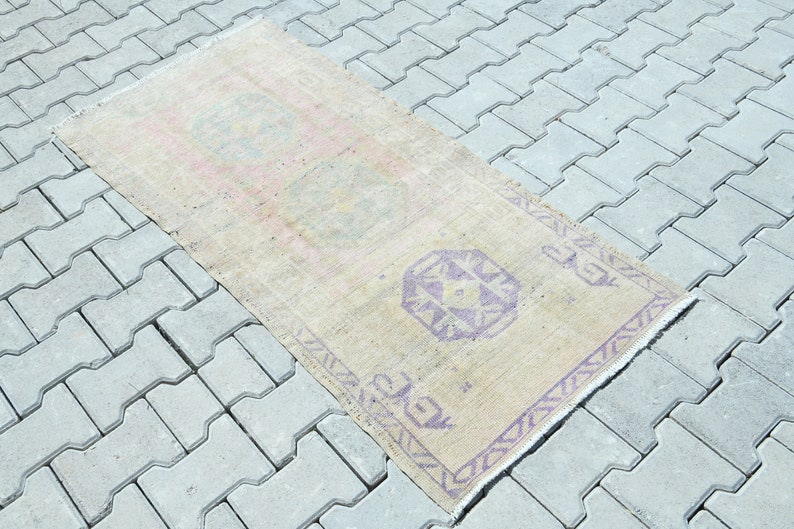 May include: A rectangular, beige rug with a faded, vintage appearance. The rug features repeating geometric patterns in shades of purple and pink. The rug is placed on a gray, concrete surface.