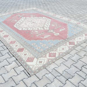 May include: A vintage rug with a pink, blue, and white geometric pattern. The rug has a faded, distressed look and is laid out on a gray concrete surface.