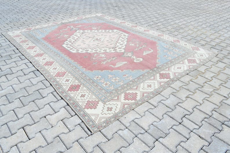 May include: A vintage rug with a pink, blue, and white geometric pattern. The rug has a faded, distressed look and is laid out on a gray concrete surface.