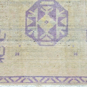 May include: Close-up of a beige rug with a central geometric design in lavender. The rug features a repeating border pattern and decorative elements in the same lavender hue. The rug has a fringe along the bottom edge.