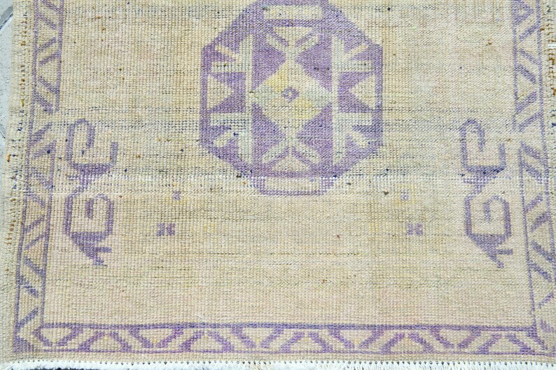 May include: Close-up of a beige rug with a central geometric design in lavender. The rug features a repeating border pattern and decorative elements in the same lavender hue. The rug has a fringe along the bottom edge.
