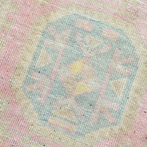 May include: Close-up of a vintage rug with a faded, textured appearance. The rug features a central geometric design in shades of blue, pink, and yellow, set against a neutral beige background. The design has a distressed, antique look.