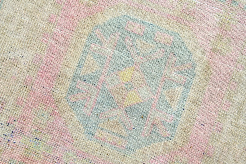 May include: Close-up of a vintage rug with a faded, textured appearance. The rug features a central geometric design in shades of blue, pink, and yellow, set against a neutral beige background. The design has a distressed, antique look.