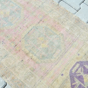 May include: A rectangular, vintage-style rug with a faded, distressed appearance. The rug features a cream-colored base with pink, blue, and purple geometric patterns. The rug is likely made of woven fabric.