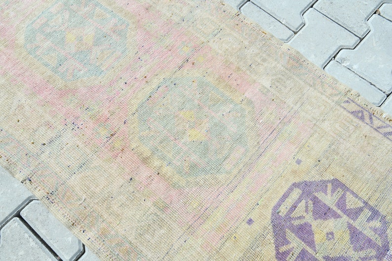 May include: A rectangular, vintage-style rug with a faded, distressed appearance. The rug features a cream-colored base with pink, blue, and purple geometric patterns. The rug is likely made of woven fabric.