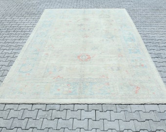 Könnte beinhalten: Ein hellbeiger Teppich mit einem verblassten Blumenmuster in Blautönen, Rosa und Orange. Der Teppich hat einen Fransenrand und liegt auf einer grauen Betonfläche.