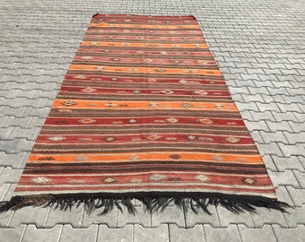 Pode incluir: Um tapete vintage tecido à mão com um padrão listrado colorido em tons de vermelho, laranja, marrom e branco. O tapete tem uma borda de franja e é feito de lã.