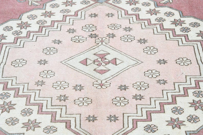 May include: A pink and white rug with a geometric pattern. The rug has a central diamond shape with a smaller diamond inside. The rug is decorated with stars and flowers.
