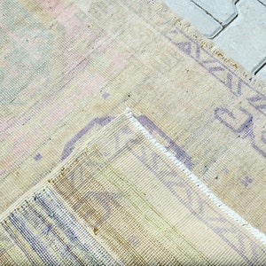 May include: A vintage-style rug with a faded, distressed appearance. The rug features a mix of pastel colors, including pink, blue, and yellow, with a beige background. The edges are trimmed with a purple geometric border.