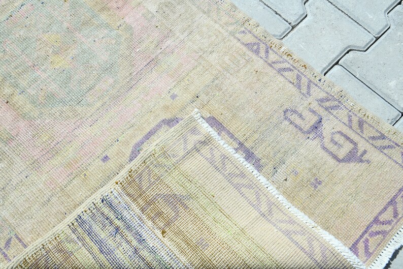 May include: A vintage-style rug with a faded, distressed appearance. The rug features a mix of pastel colors, including pink, blue, and yellow, with a beige background. The edges are trimmed with a purple geometric border.