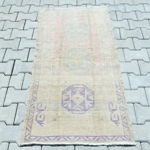 May include: A rectangular, vintage-style rug with a faded, distressed appearance. The rug features a beige background with a central geometric design in shades of purple and teal. The rug's dimensions are approximately 2 x 4 feet.