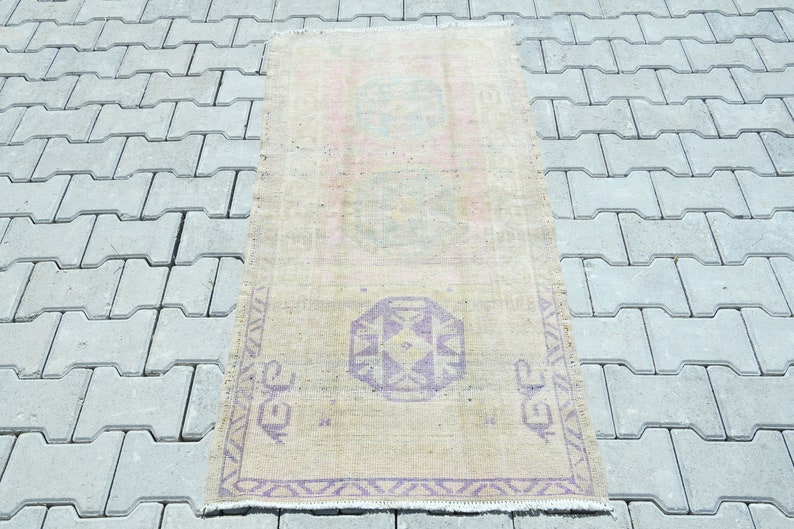 May include: A rectangular, vintage-style rug with a faded, distressed appearance. The rug features a beige background with a central geometric design in shades of purple and teal. The rug's dimensions are approximately 2 x 4 feet.