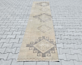May include: A beige and brown runner rug with a geometric pattern. The rug has a diamond shape in the center with a smaller diamond inside. The rug is on a gray concrete surface.