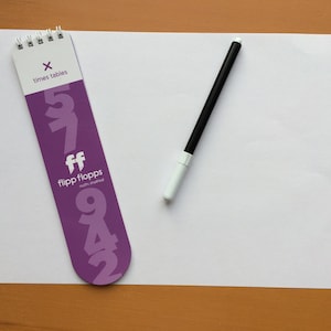 May include: Purple times tables flip card with the numbers 5, 7, 9, 4, and 2 printed on it. The card has the text "flip flops math inspired" printed on it. A black marker with a white cap is on a white sheet of paper.