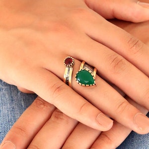 May include: Two rings with gemstone accents. One ring features a small, round, red gemstone. The other ring has a pear-shaped, green gemstone. Both rings have gold-colored bands and are displayed on a hand.