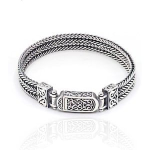 May include: Silver bracelet with a woven design. The bracelet has an ornate clasp with decorative patterns. The metal bracelet is displayed on a white background. A brown tag with text is also visible.