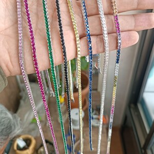 May include: A collection of tennis bracelets in various colors, including pink, magenta, green, black, yellow, blue, and a gradient of colors. Each bracelet features a row of gemstones set in a silver metal. The bracelets are displayed against a neutral background.