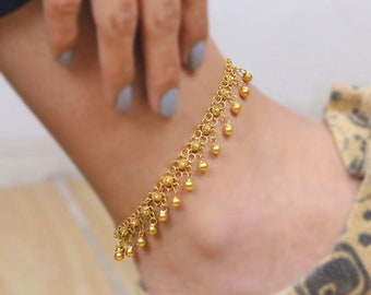 May include: A gold anklet with a delicate chain and dangling beads. The anklet is worn on a person's ankle.