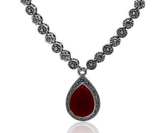 May include: Silver necklace with a teardrop-shaped, deep red pendant. The pendant is framed with small, dark stones. The necklace chain features a floral design. The necklace is displayed on a white background.