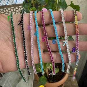 May include: Eight silver tennis bracelets with different colored stones. The bracelets are lined up in a row, with the stones facing the viewer. The stones are green, black, pink, turquoise, red, blue, clear, and rainbow.