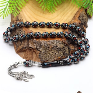 Puede incluir: Cuentas de oración negras con detalles azules y rojos, detalles plateados y un borla plateada. Las cuentas están sobre una superficie de madera. La imagen incluye el texto "Premium Ahşap Kutu Hediye".