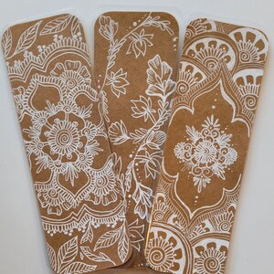 May include: Three brown paper bookmarks with white intricate designs. The designs are different on each bookmark and feature floral and geometric patterns.