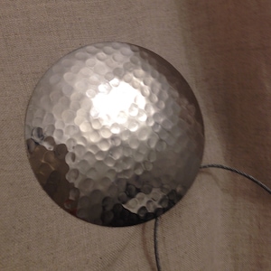 May include: A silver, hammered metal disc with a cord attached. The disc has a textured, hammered surface.