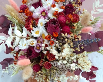 May include: A vibrant bouquet of dried flowers in a brown, shallow bowl. The arrangement features a mix of textures and colors, including deep red roses, white daisies, orange accents, and fluffy pink elements. The overall aesthetic is rustic and natural.