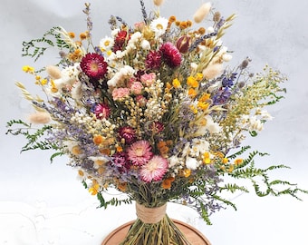 Könnte beinhalten: Ein Trockenblumenstrauß mit einer Vielzahl von Farben, darunter Weiß, Gelb, Lila, Rosa und Rot. Der Strauß ist mit Schnur gebunden und liegt auf einer Holzoberfläche.