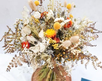 May include: A dried flower bouquet with a mix of white, yellow, orange, and brown flowers. The bouquet is tied together with a brown ribbon and is sitting on a wooden surface.