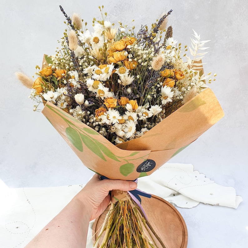 May include: A bouquet of dried flowers in shades of yellow and white, wrapped in brown paper with green leaf designs. The bouquet includes dried roses, daisies, and other wildflowers.