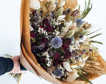 May include: A bouquet of dried flowers wrapped in brown paper. The bouquet includes burgundy roses, blue thistle, white baby's breath, and dried grasses.
