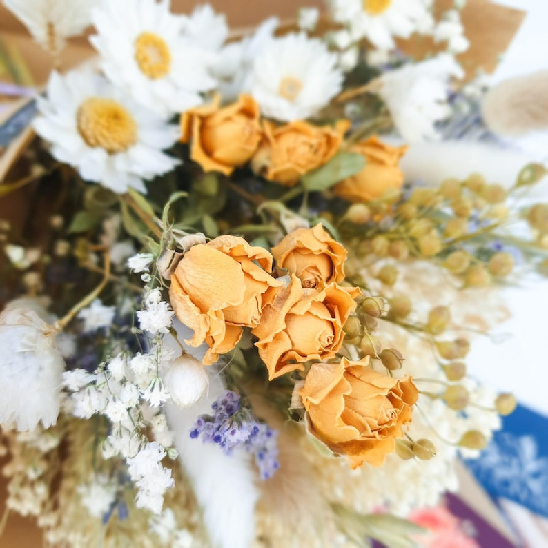 May include: A bouquet of dried flowers with white daisies, dried grasses, and orange roses.