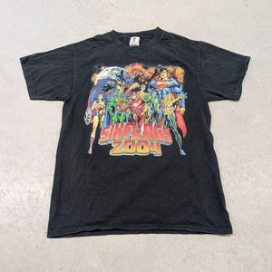 Puede incluir: Una camiseta negra de manga corta con un estampado gráfico colorido de superhéroes de DC Comics, incluyendo a Superman, Batman y Wonder Woman. El texto "SIX FLAGS 2004" está impreso en letras grandes rojas y amarillas en el centro del pecho.