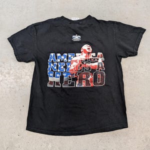 Puede incluir: Camiseta negra con el texto "AMERICA NEEDS A HERO" en rojo, blanco y azul. El gráfico presenta a una persona con los brazos levantados. La camiseta tiene cuello redondo y mangas cortas.