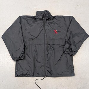 Könnte beinhalten: Schwarze Windjacke mit durchgehendem Reißverschluss, hohem Kragen und Kordelzug. Auf der Brust befindet sich ein rotes, gesticktes "X"-Logo. Die Jacke hat lange Ärmel und eine lockere Passform.