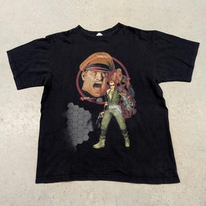 May include: Black t-shirt with a graphic of a man in a military hat and a man with sunglasses and a gun. The graphic is in shades of red, brown, green, and grey. The shirt is made of a soft material.