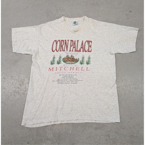 Könnte beinhalten: Hellgraues T-Shirt mit dem roten Schriftzug "Corn Palace" und "Mitchell South Dakota" darunter. Das T-Shirt hat eine Grafik eines Gebäudes und Text, der schwer zu lesen ist.