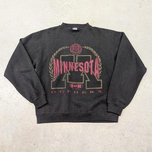 May include: Dark gray sweatshirt with a crew neck. The front features a maroon and gold design with the word "MINNESOTA" in large letters, along with "U of M" and "GOPHERS". The design is framed by a gold laurel wreath.