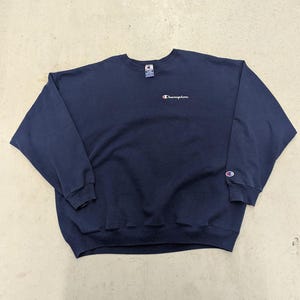 Puede incluir: Sudadera Champion azul marino. La sudadera de cuello redondo tiene el logotipo de Champion bordado en blanco y rojo en el pecho y en el puño izquierdo de la manga. La etiqueta Champion es visible en la parte posterior del cuello.